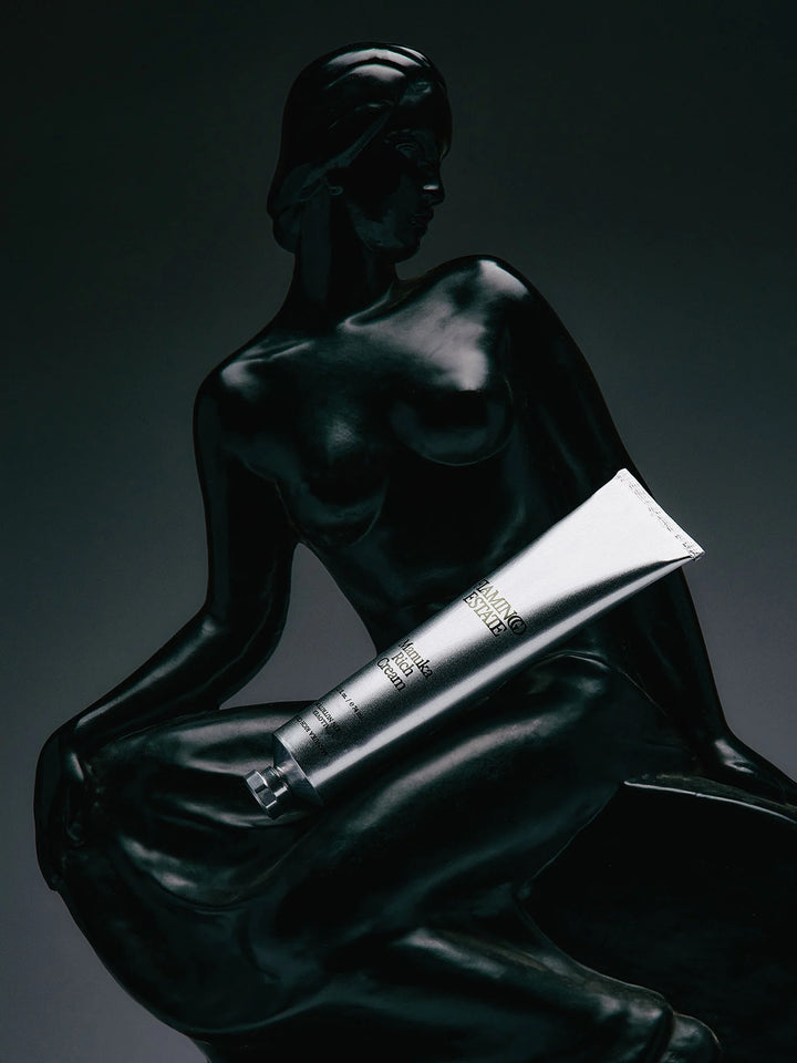 Silver aluminum cream tube resting against a dark bronze sculpture of a seated figure