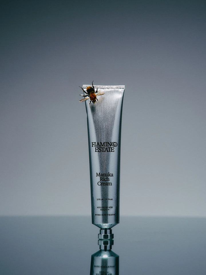 Silver aluminum cream tube standing upright on a grey surface with a honeybee resting on its crimped cap
