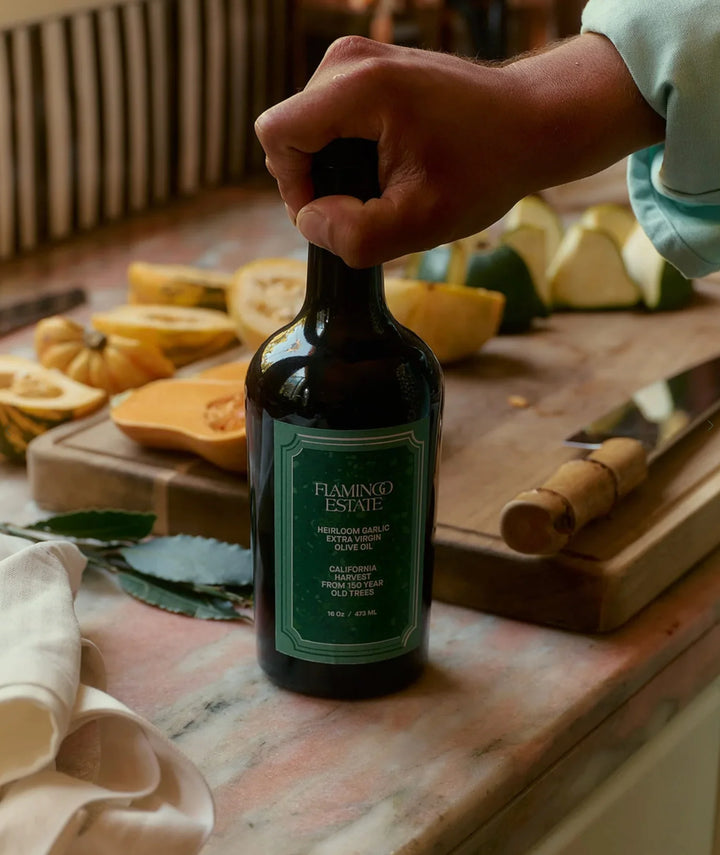 A hand lifting the bottle by its cap from a marble counter, with halved squash on a wooden board behind