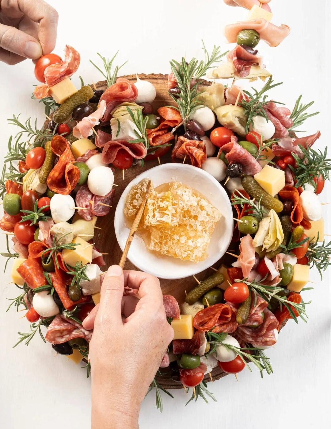 Hands serving from a wreath-shaped charcuterie board with a small bowl of honeycomb at the center