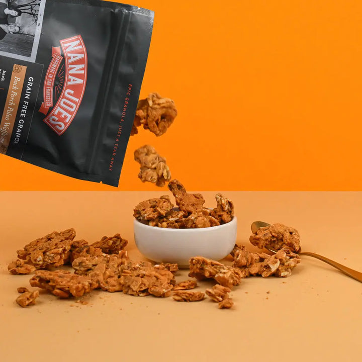 Granola clusters pouring from a Nana Joes bag into a white bowl against an orange background