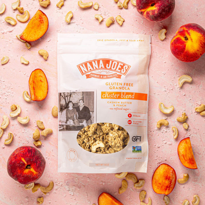Nana Joes Cashew Butter and Peach cluster blend granola bag on a pink surface with fresh peach slices and cashews