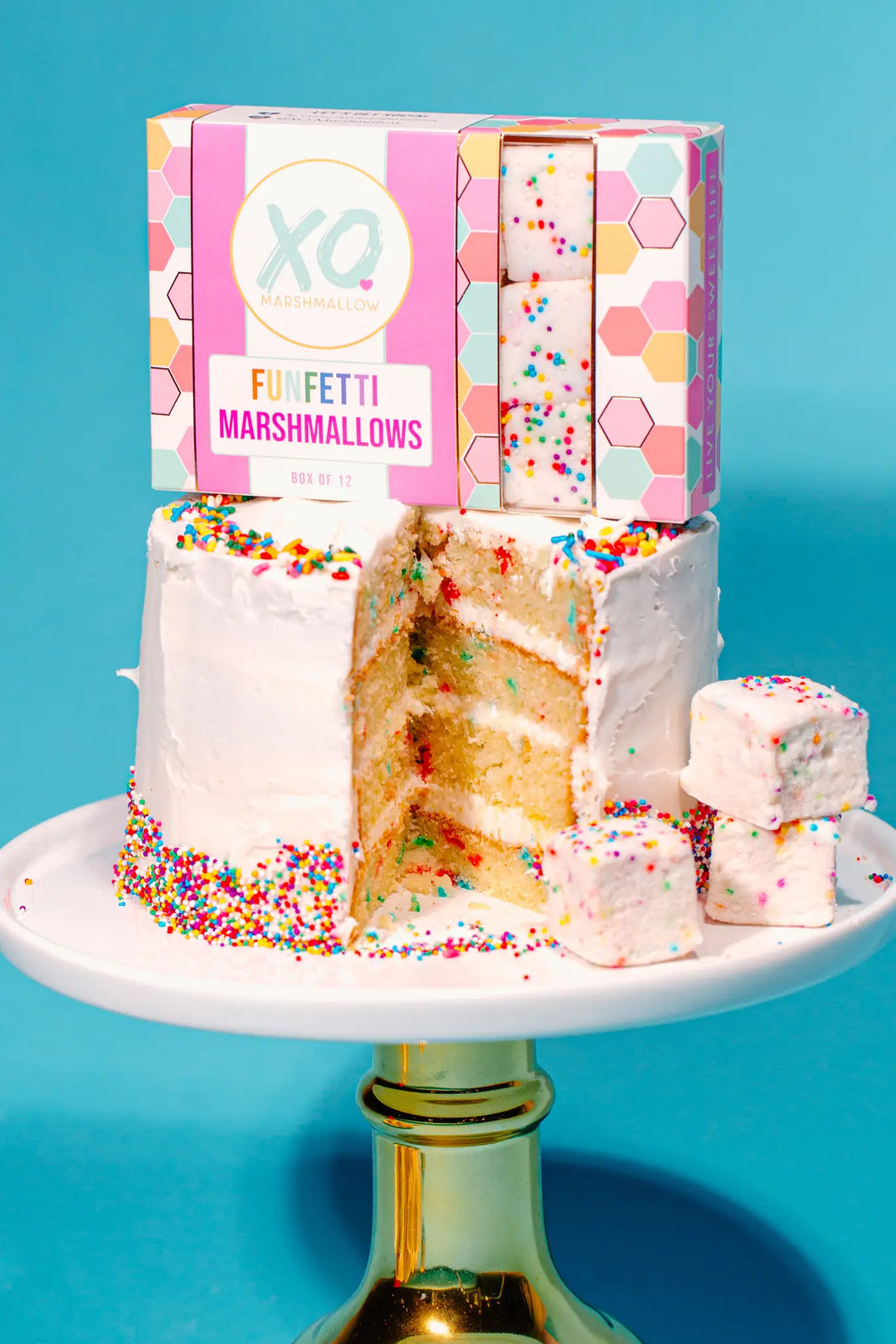 Box of XO Funfetti marshmallows on top of a layered sprinkle cake, with marshmallow cubes beside it on a gold cake stand
