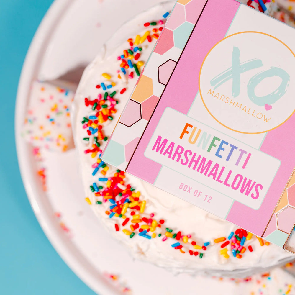 Close view of the XO Funfetti Marshmallows box of 12 resting on a frosted cake edged with rainbow sprinkles