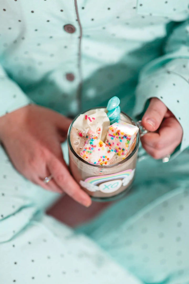 Person in pajamas holding a glass mug of hot cocoa topped with sprinkle marshmallows
