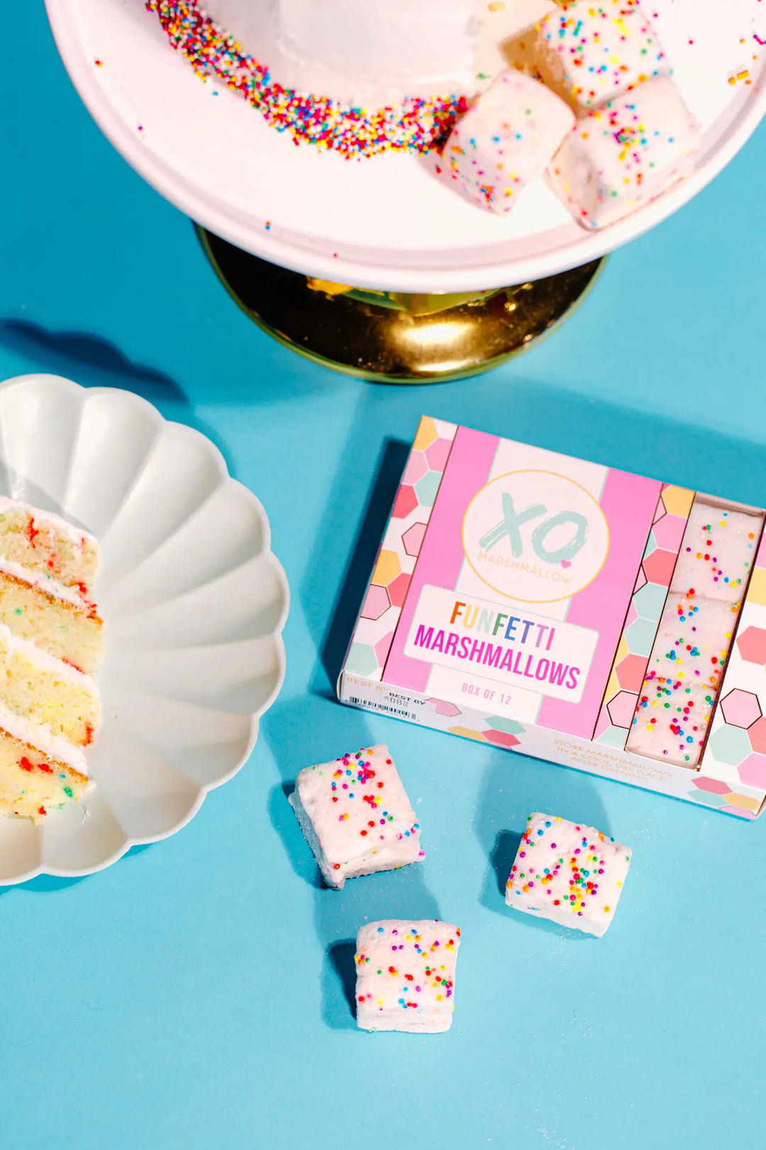 XO Funfetti marshmallow box on a blue surface with loose marshmallows, a slice of cake and a cake stand