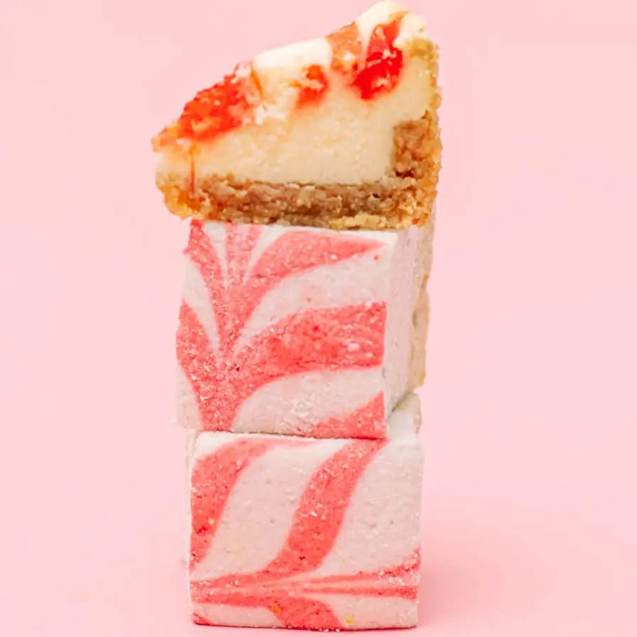 Two swirled marshmallows stacked under a small slice of strawberry cheesecake on a pink background