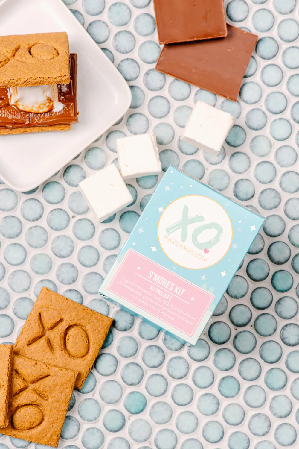 Flat lay of an assembled s'more, marshmallows, chocolate pieces and XO-stamped graham crackers around the kit box on blue penny tile