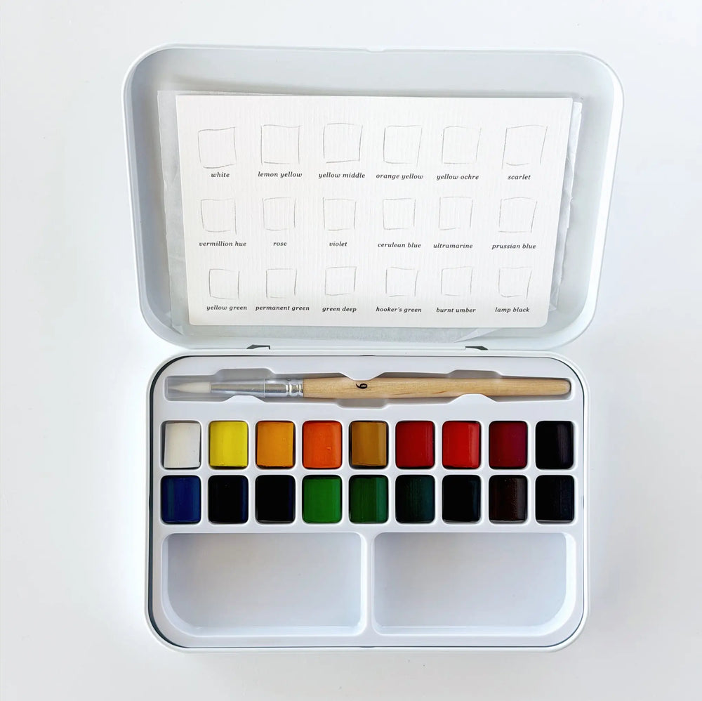 Open white metal watercolor tin with 18 paint pans in two rows, a wooden size 6 round brush, and a color swatch card inside the lid