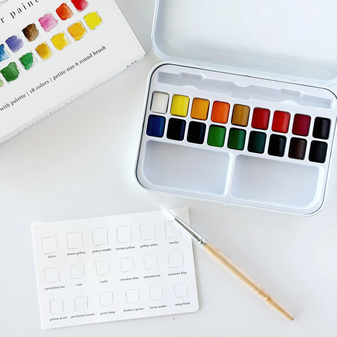 Open watercolor tin beside its box, with the swatch card and size 6 round wooden brush laid out on a white table