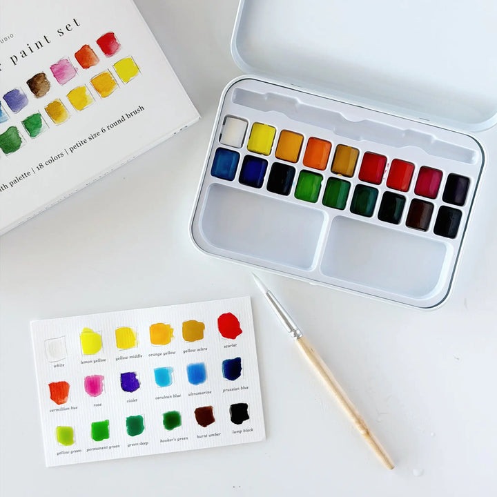 Flat lay of the open watercolor tin, box, partially painted swatch card, and wooden round brush