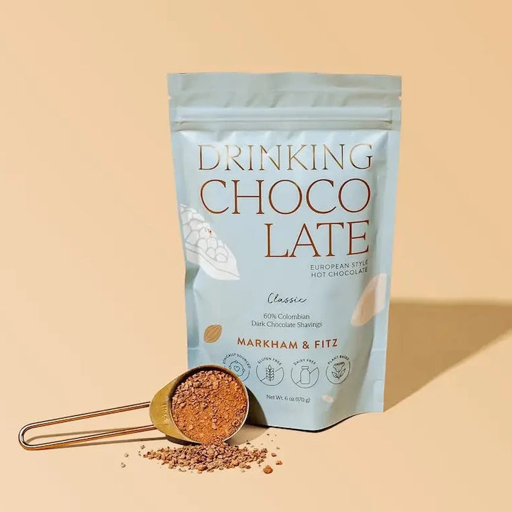 Pale blue matte pouch of Markham &amp; Fitz Classic drinking chocolate beside a copper scoop heaped with dark chocolate shavings