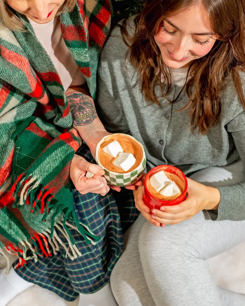 Two people wrapped in plaid blankets holding ceramic mugs of hot chocolate topped with marshmallows
