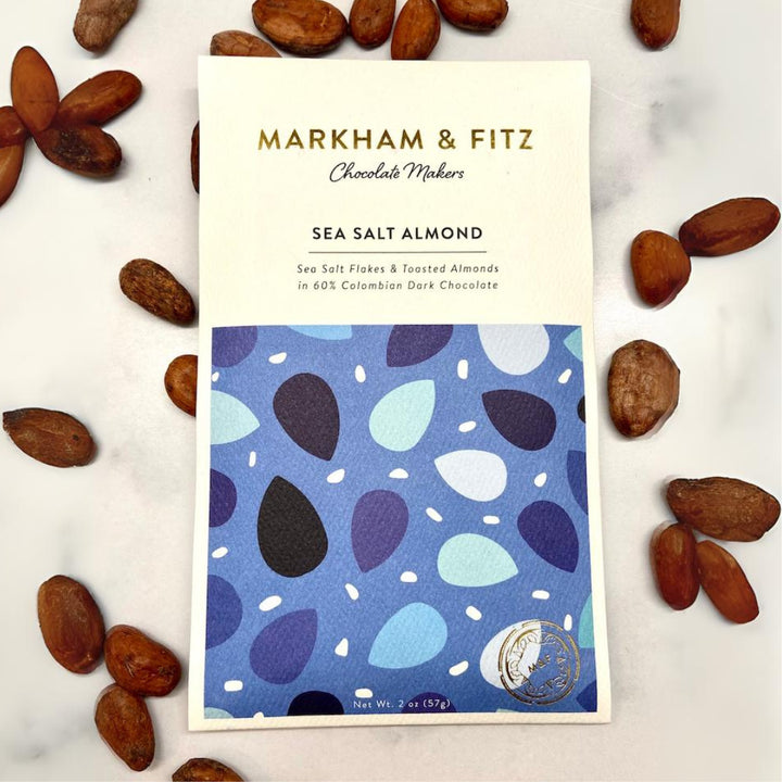 Markham &amp; Fitz Sea Salt Almond bar in a cream and blue sleeve on white marble, surrounded by whole cocoa beans