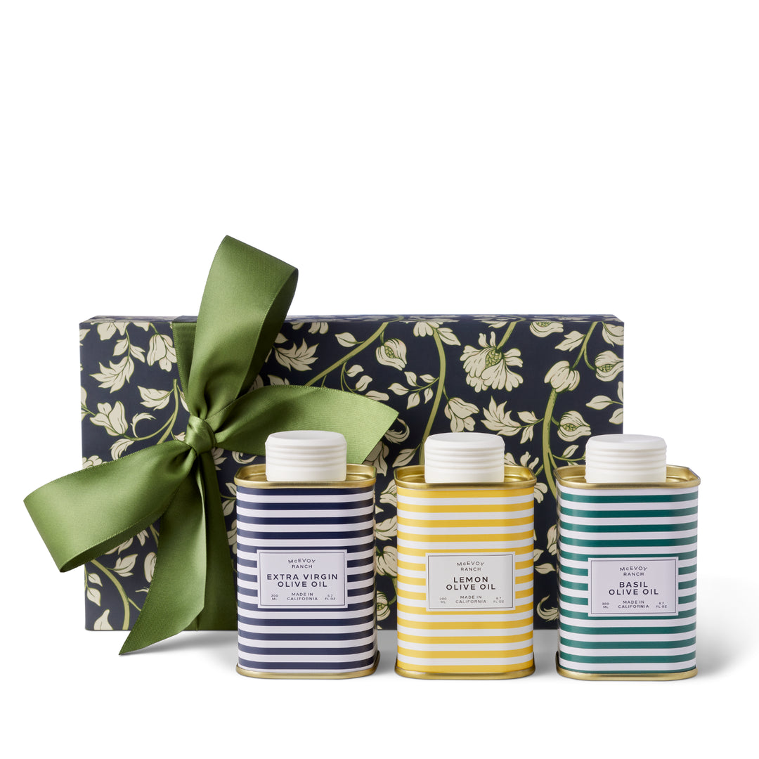 Navy floral McEvoy Ranch gift box with a green satin ribbon, with three striped olive oil tins in navy, yellow and green lined up in front