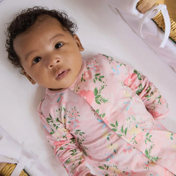 Baby in floral pink footie sleeper lying in a woven bassinet with white bedding