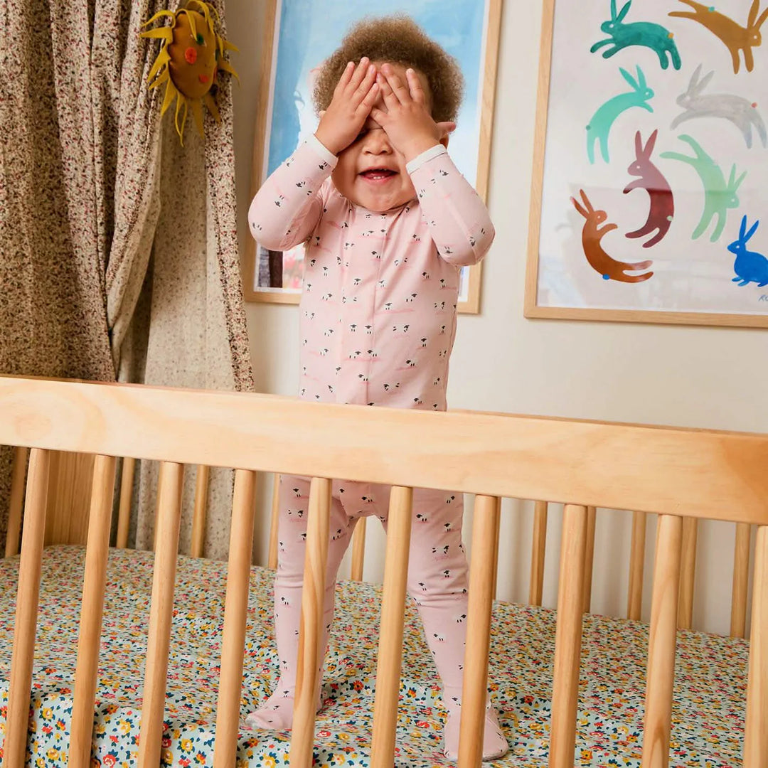 Toddler in soft pink baby footie pajamas playing in a natural wood crib