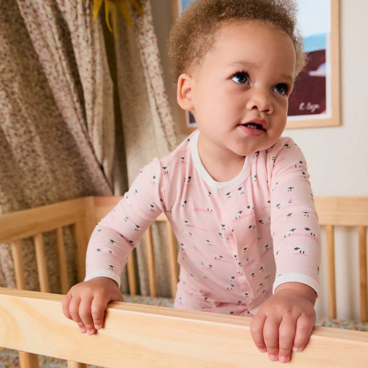 Baby in a pink footed sleeper with sheep print, standing inside a wooden crib