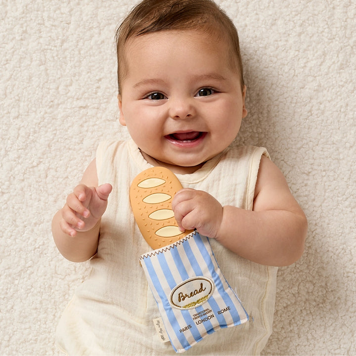 Smiling baby holding baguette-shaped teether in striped fabric pouch on soft cream blanket
