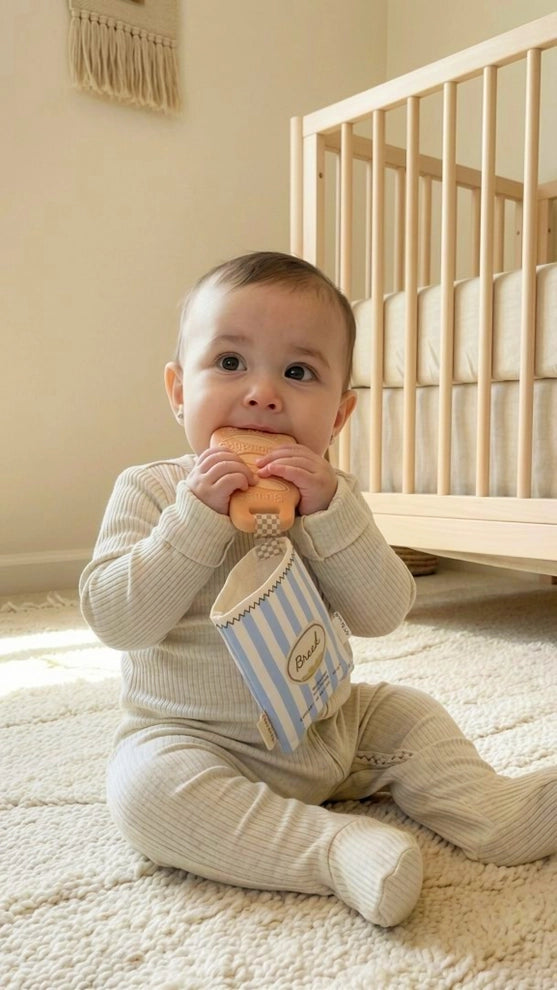 Baby in neutral pajamas chewing on a striped teether toy, sitting on cream rug beside crib