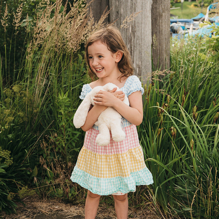 A young girl stands outdoors in a garden setting, smiling and holding a white stuffed bunny. She is wearing a pastel gingham dress with light blue, pink, and yellow checks, and white slide sandals.