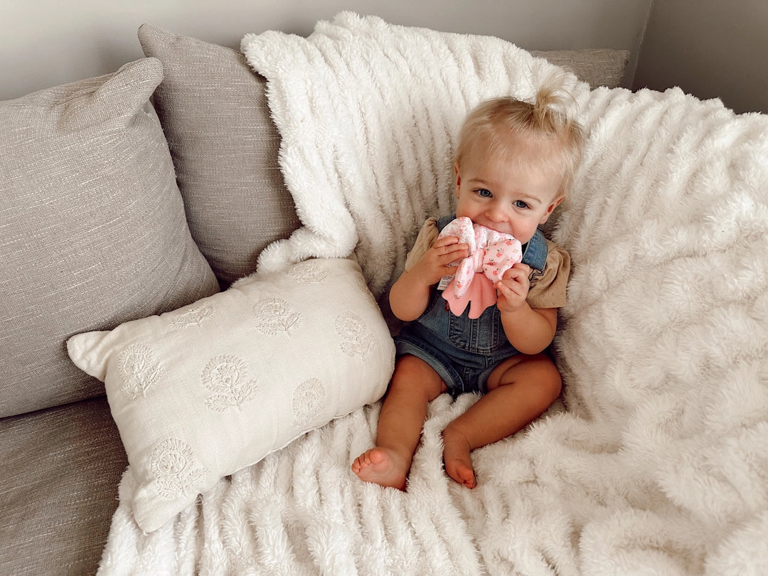Baby holding pink bow-shaped teether on cream textured blanket and neutral pillows