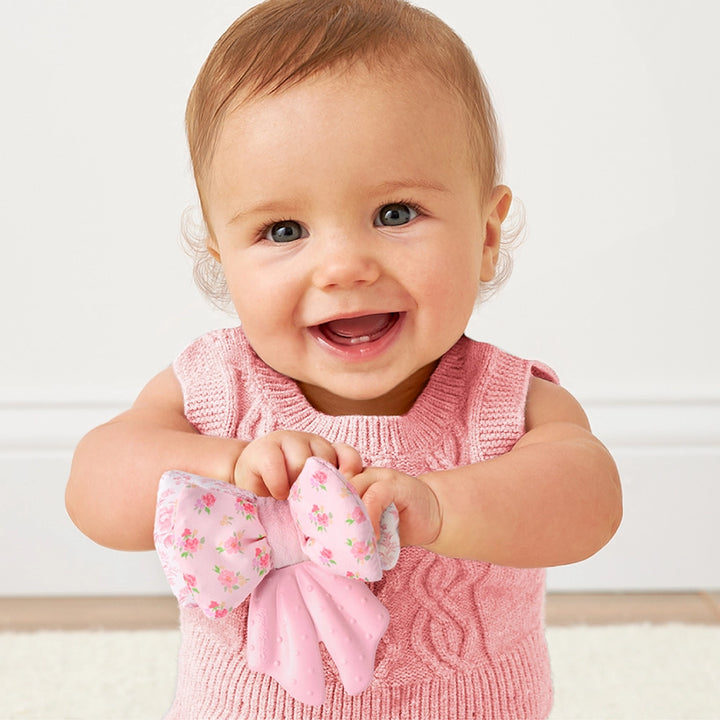 Smiling baby in pink sweater holding a floral pink bow-shaped teether toy