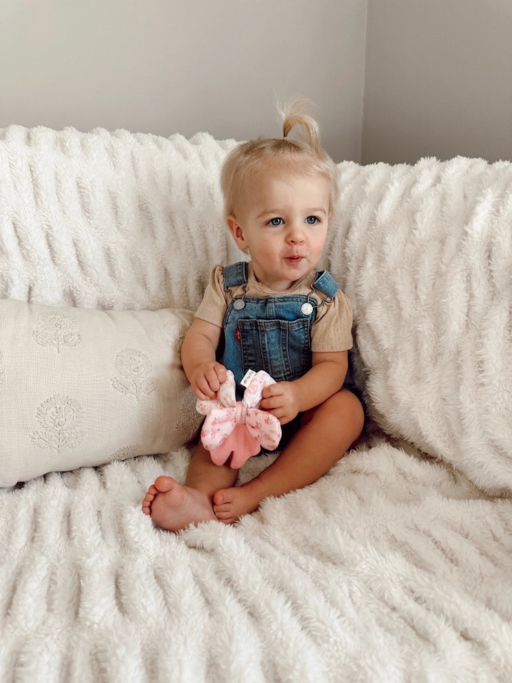 Baby holding pink bow-shaped crinkle teether on white textured blanket