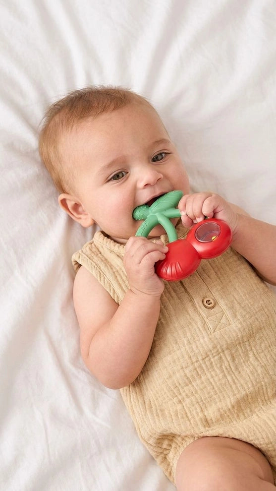 Baby holding red cherry-shaped teether toy while lying on white blanket