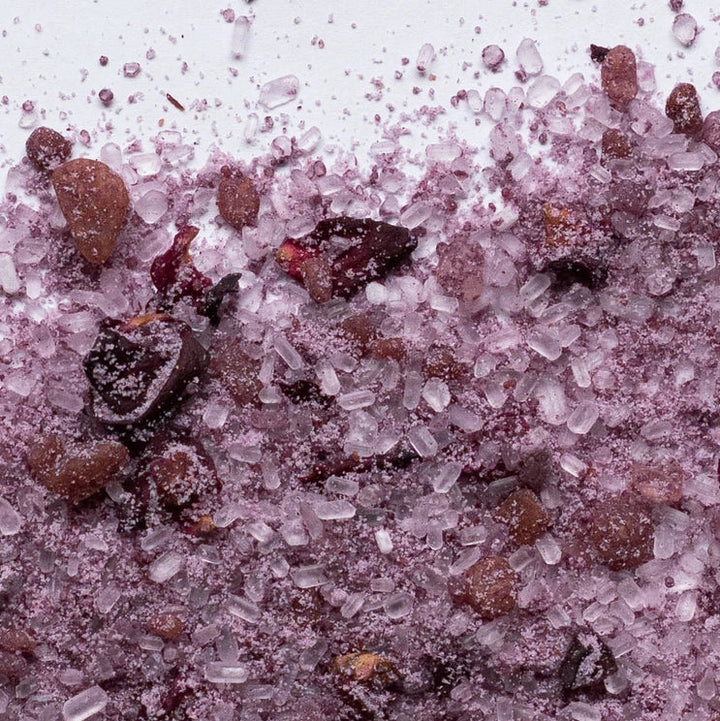 Closeup of pink bath soak crystals with dried botanicals on a white surface