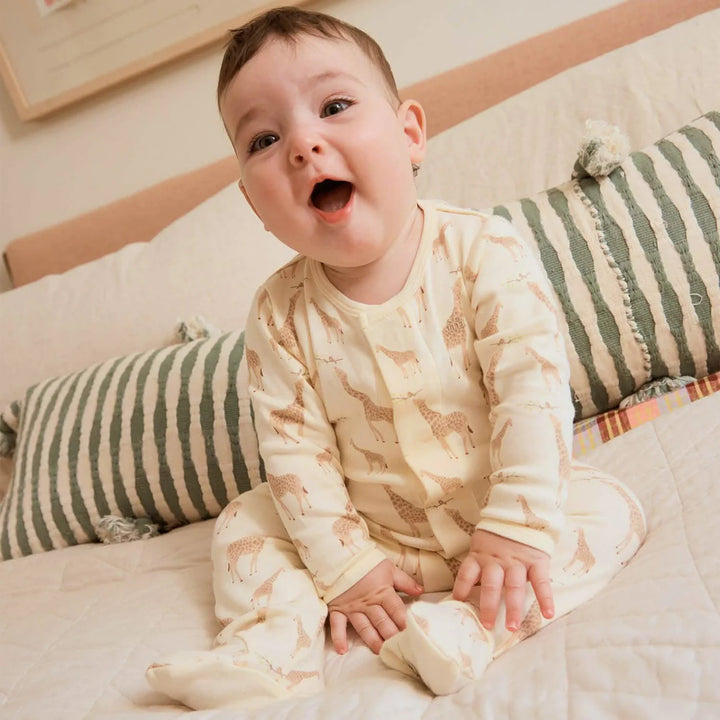 Baby wearing cream footie pajamas with giraffe print, sitting on a bed