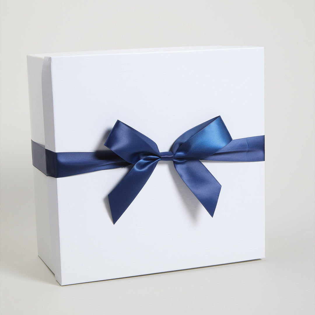 A white square gift box tied with a wide navy blue satin ribbon and bow, photographed on a clean light beige studio background.