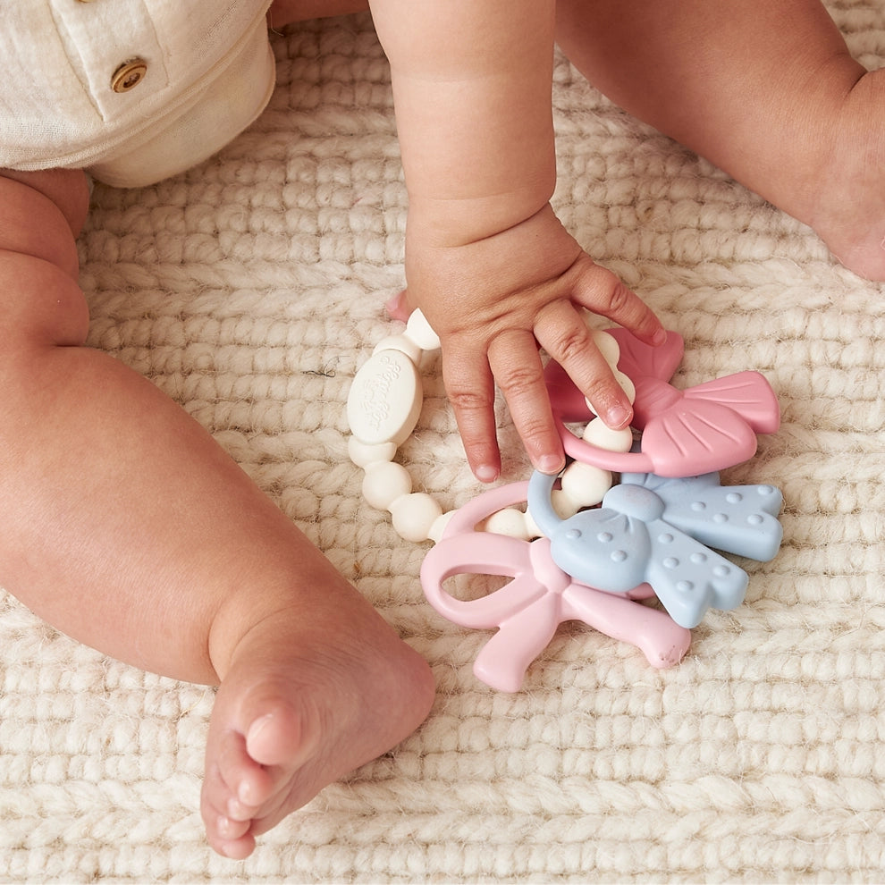 Baby holding pastel silicone teething toy with blue and pink elements on cream knit rug