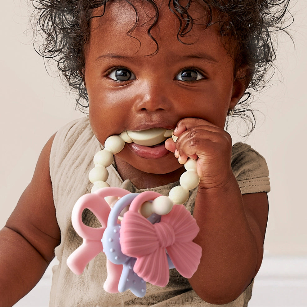 Baby holding pastel silicone teething toy with beaded handle and bow-shaped charms