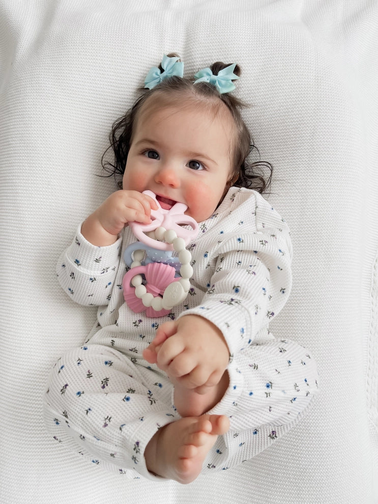 Baby holding a pastel silicone teething toy, wearing floral pajamas on a white blanket