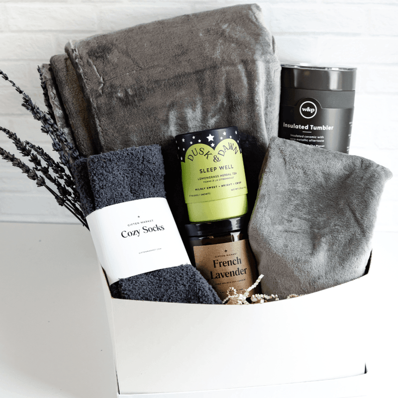 A light gray gift box opened to display its contents, which include cozy socks, a fleece neck wrap, an insulated ceramic tumbler, a soy wax candle, and other items.