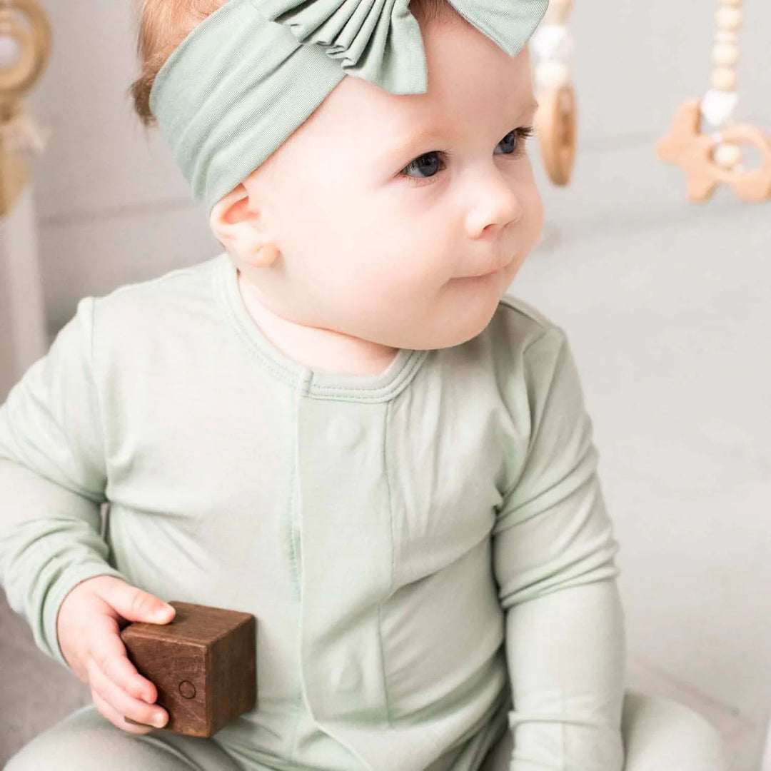 Baby in sage green footie pajamas with a matching headband holding a wooden block