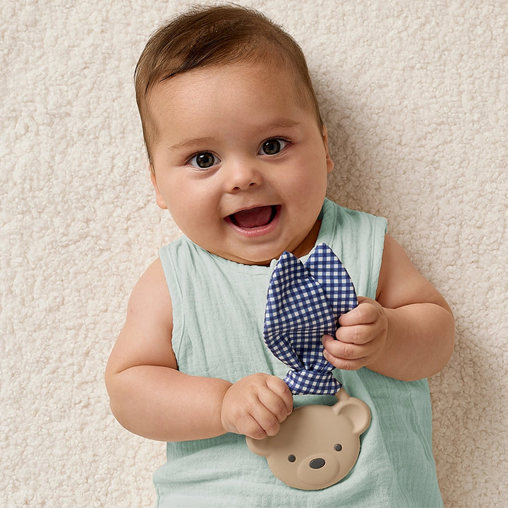 Baby holding taupe bear teether with blue gingham fabric on soft textured rug