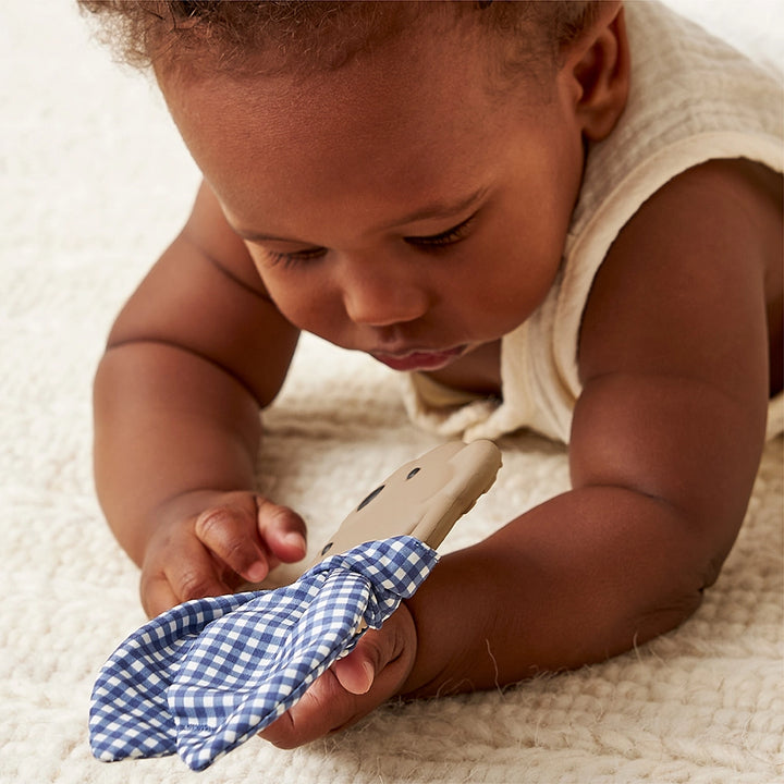 Baby holding taupe bear-shaped teether with blue gingham fabric on a cream blanket