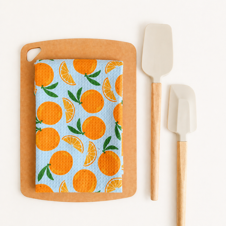 Wooden cutting board with a towel featuring orange pattern, accompanied by white spatulas on a white background.
