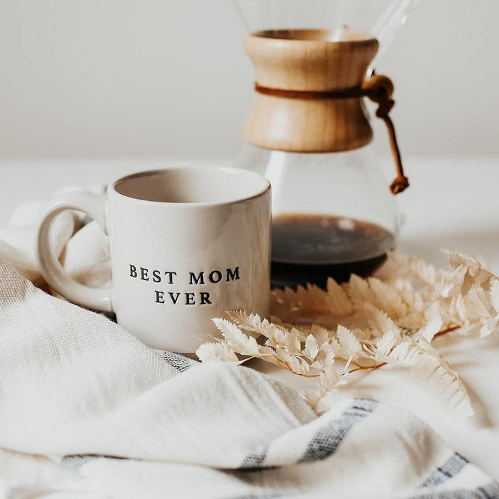 Stoneware mug with 'Best Mom Ever' text, beside coffee maker, on neutral fabric