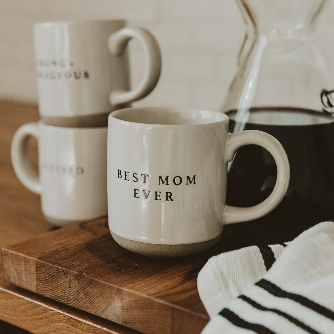 Stoneware coffee mug with 'Best Mom Ever' text on wooden table, two mugs and carafe in background