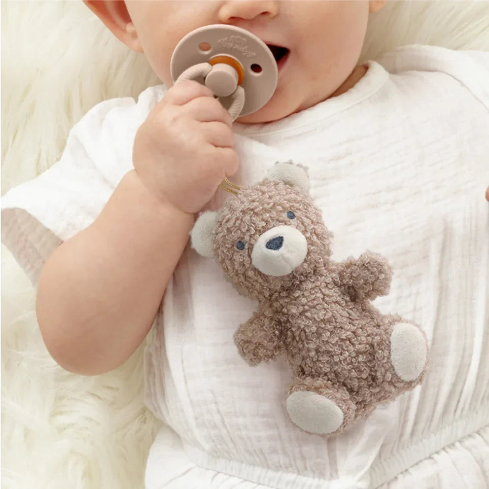 Baby wearing white holding natural rubber pacifier with plush bear toy attached