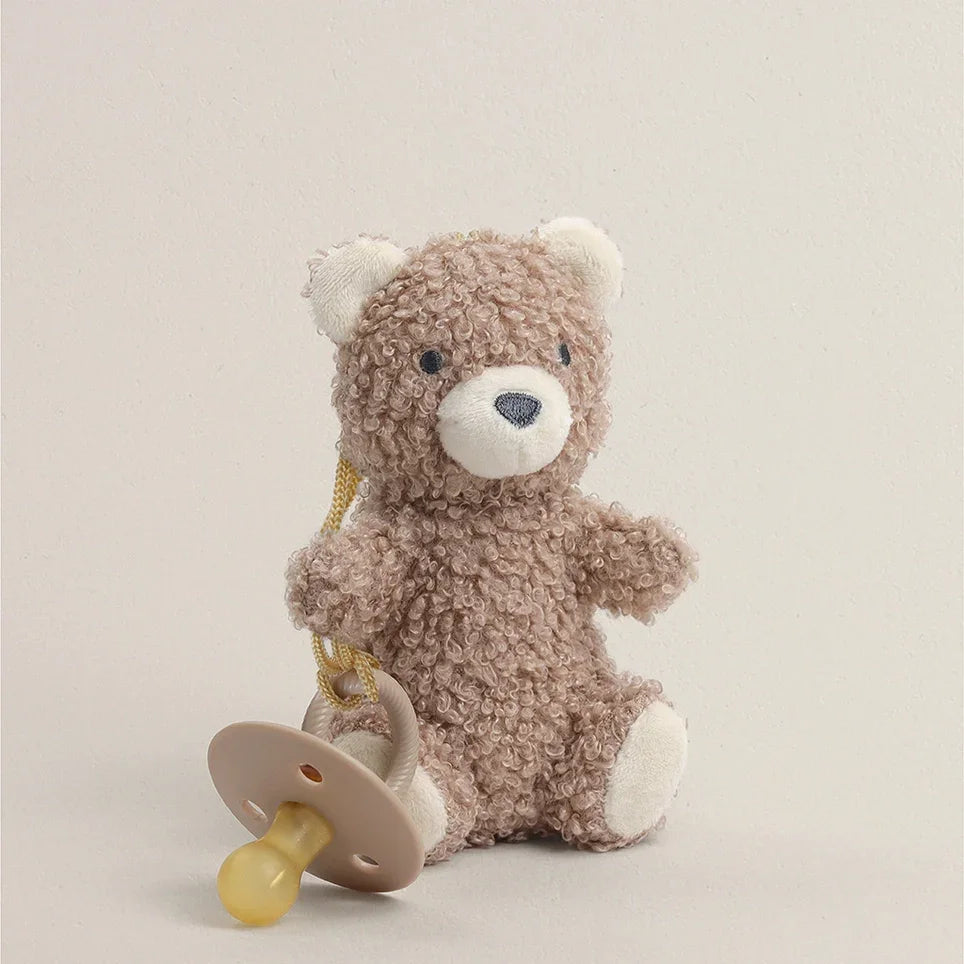 Plush teddy bear holding a natural rubber pacifier against a neutral background
