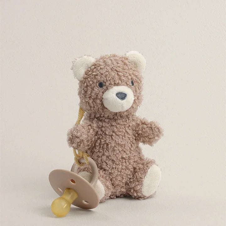 Plush teddy bear holding a natural rubber pacifier against a neutral background