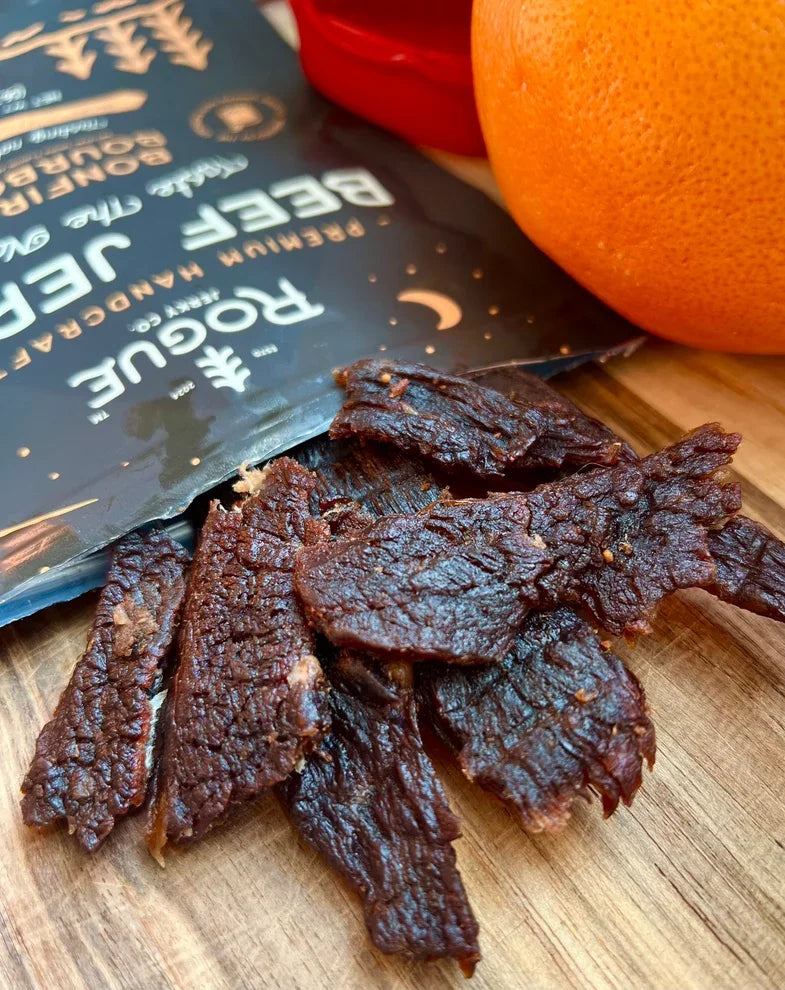 Gourmet beef jerky slices on a wooden board beside unopened snack packaging and an orange