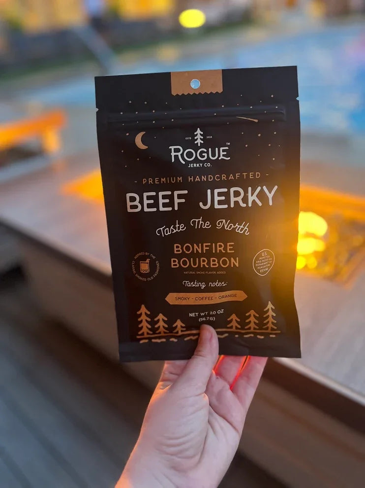 Hand holding a premium black pouch of bonfire bourbon beef jerky outdoors by a fire.