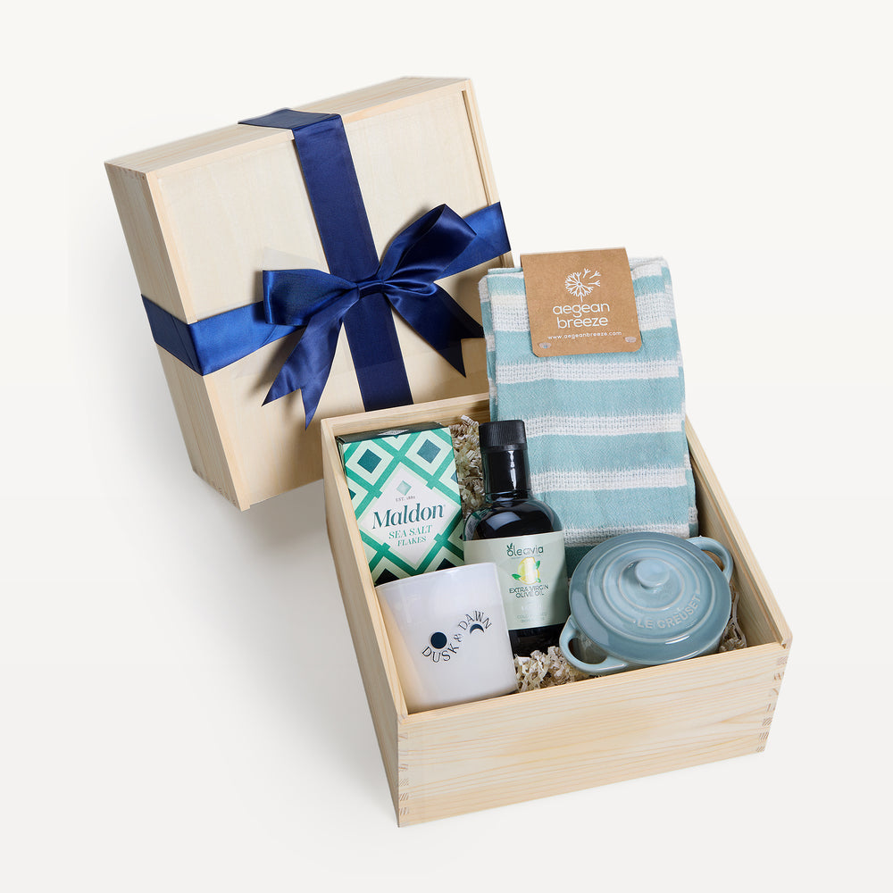 Coastal Kitchen crate open in front, bow-tied lid behind. Aegean Breeze striped tea towel, Maldon sea salt, olive oil bottle, Dusk & Dawn candle, sage-blue Le Creuset mini cocotte. Giften Market.