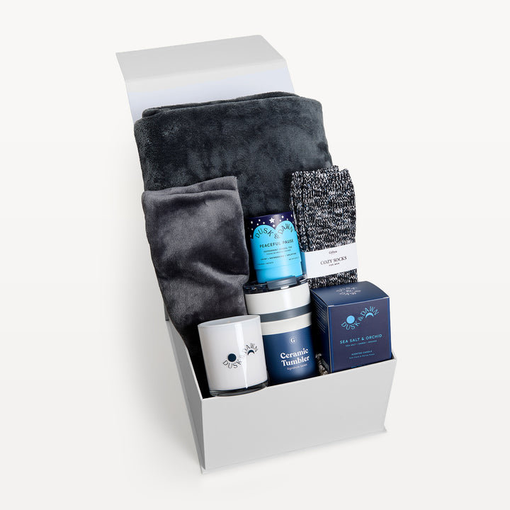 Comfort &amp; Care Light Gray, white gift box, lid open. Charcoal faux-fur throw, gray Cozy Socks, Feel Well tea, Dusk &amp; Dawn Lavender candle box, cream candle, navy Ceramic Tumbler. Giften Market.