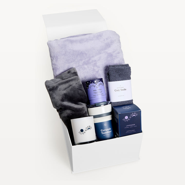 Comfort &amp; Care with the Lavender blanket, white gift box, lid open. Lilac faux-fur throw behind a charcoal throw, gray Cozy Socks, Feel Well tea, Lavender candle, Ceramic Tumbler. Giften Market.